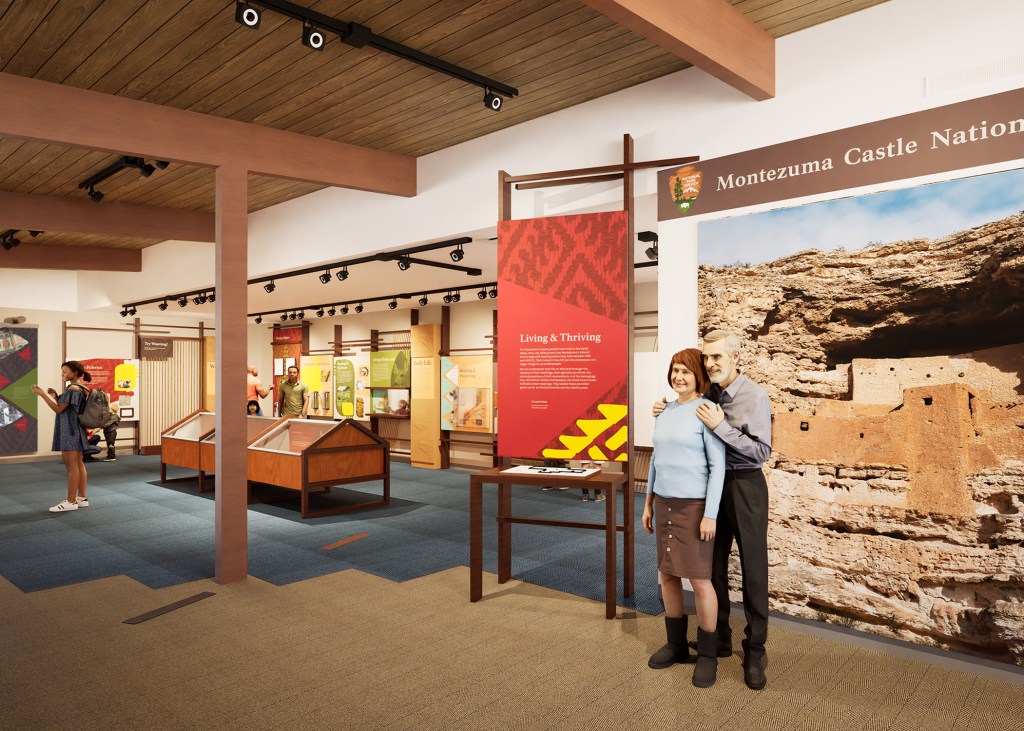 Design rendering of visitor center displays at Montezuma Castle National Monument showing the approach designers took to accessible graphics. Track lighting mounted on the wooden ceiling ensures the space is well lit. The foreground features two figures posing in front of a large photographic backdrop showcasing the ancient cliff dwellings, providing a sense of scale. Several display cases and information panels are visible throughout the space. A rendering of a young woman in a blue dress in the background enhances the sense of depth and scale.