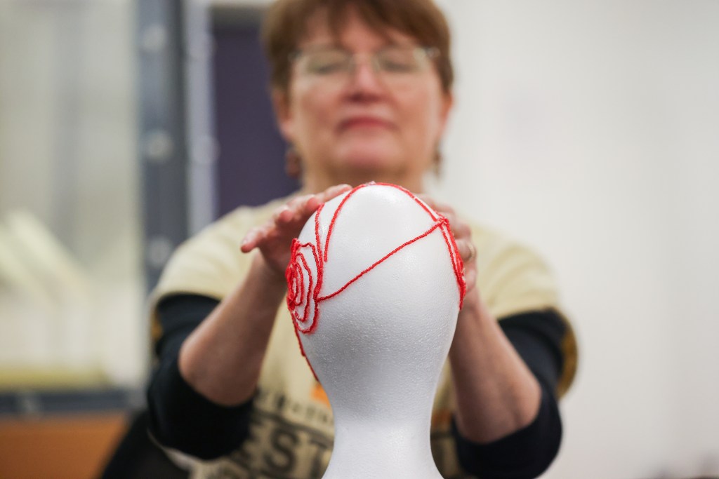 A woman with short brown hair and glasses is touching a Styrofoam mannequin head. The head is adorned with bright red pipe cleaners that have been arranged is a specific pattern. The image captures only the back of the mannequin's head; from the placement of the woman's hands, it seems likely that the pattern continues around the head to the figure's face.