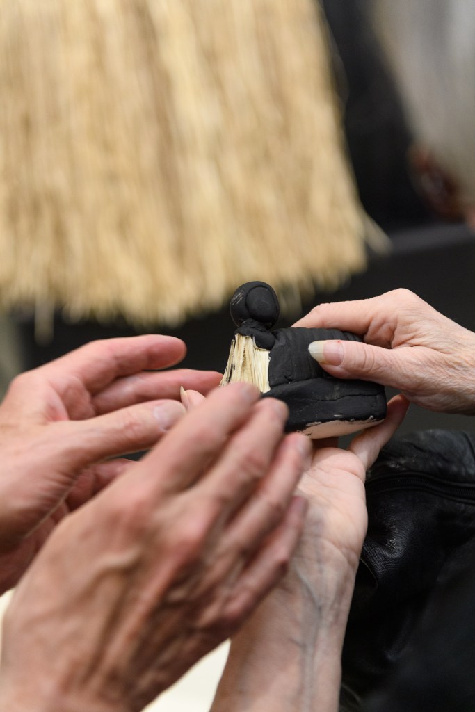 A close-up image of participants in a Mind's Eye program at the Solomon R. Guggenheim Museum touching a replica of an artwork by Simone Leigh. The replica is black, with a rounded head and body, with a skirt of straw extending from the lower part. It is small enough to be held comfortably in the palm a hand. One set of hands with long nails holds the figure while another left-reaching pair of hand touching the straw. In the background, in soft focus, a portion of Leigh's original, much larger artwork is visible.