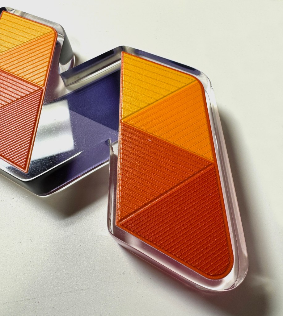 A closely cropped image of a wide and thick clear acrylic capital M, the size of a standard book, constructed from 13 purple and orange equilateral triangles. The triangles have a slightly textured hashed line on the surface that increase in density as the colors increase in saturation. 