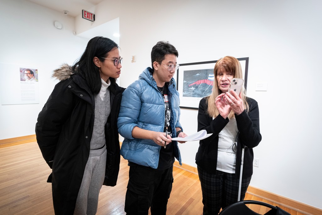 Three figures stand, conversing in a gallery space with wood floors, white walls, and a few visible 2D artworks. The woman on the right has blonde hair and a mobility cane dangling by its strap from her left arm. She is holding up a cell phone, demonstrating something on it to her two companions, who are younger than she is and dressed for winter weather.

