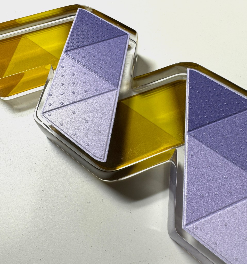 A closely cropped image of a wide and thick clear acrylic capital M, the size of a standard book, constructed from 13 purple and yellow equilateral triangles. The triangles have a slightly textured dot pattern on the surface that increase in density as the colors increase in saturation. 