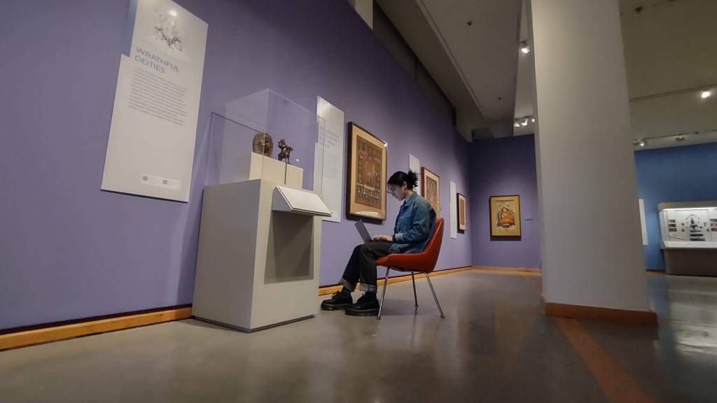 A female college student with dark hair sits in a red chair in a museum gallery filled with works of Himalayan art. She sits in front of a case holding two small sculptures that appear to be made of bronze. She is bent over her laptop typing an audio description. To hear the description, click the link in the figure caption.