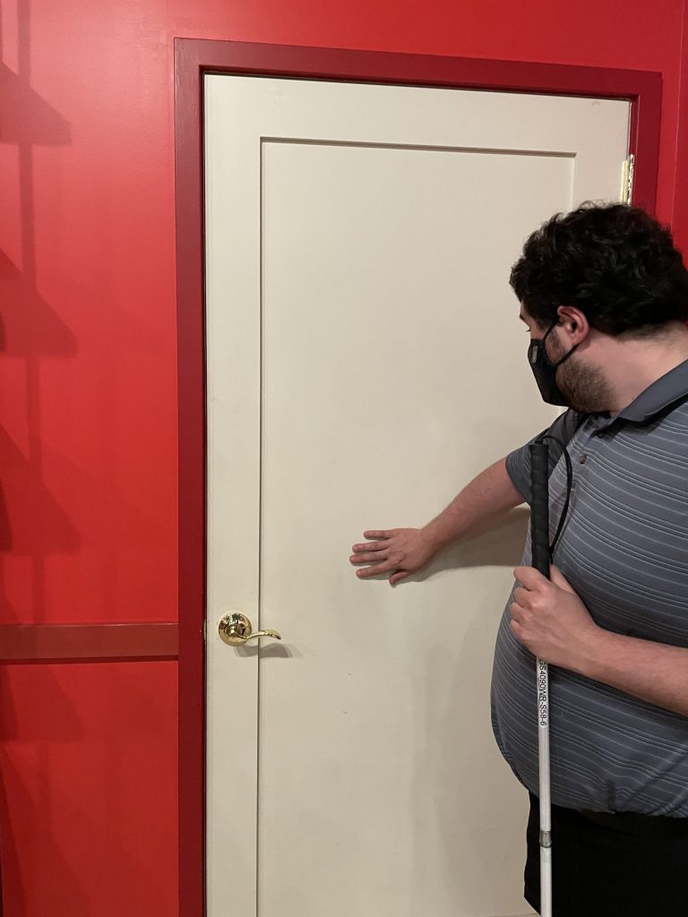 A photo of a Persian man reaching out with his right hand to touch a closed, plain beige door with a gold handle in front of him. The door is set within a bright red wall with a red frame around it, creating a high-contrast visual boundary. The man is wearing a black face mask and a gray, striped polo shirt. He is holding a white cane in his left hand. 