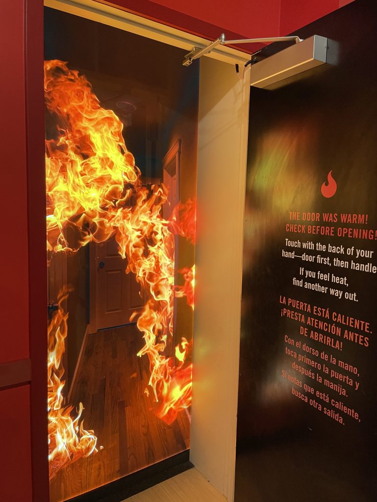 A photo of a propped open door revealing a vivid, realistic-looking backdrop image of a hallway engulfed in flames. On the backside of the open door is a bilingual fire safety warning in English and Spanish, printed on a dark wall panel. The English text reads, “THE DOOR WAS WARM! CHECK BEFORE OPENING! Touch with the back of your hand—door first, then handle. If you feel heat, find another way out.” The Spanish translation is below.