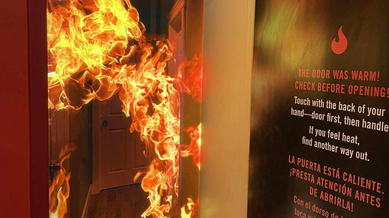 A photo of a propped open door revealing a vivid, realistic-looking backdrop image of a hallway engulfed in flames. On the backside of the open door is a bilingual fire safety warning in English and Spanish, printed on a dark wall panel. The English text reads, “THE DOOR WAS WARM! CHECK BEFORE OPENING! Touch with the back of your hand—door first, then handle. If you feel heat, find another way out.” The Spanish translation is below.