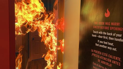 A photo of a propped open door revealing a vivid, realistic-looking backdrop image of a hallway engulfed in flames. On the backside of the open door is a bilingual fire safety warning in English and Spanish, printed on a dark wall panel. The English text reads, “THE DOOR WAS WARM! CHECK BEFORE OPENING! Touch with the back of your hand—door first, then handle. If you feel heat, find another way out.” The Spanish translation is below.