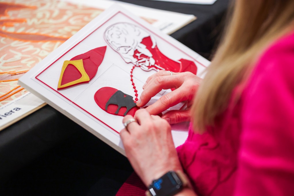 A close-up of a woman with long blonde hair wearing a pink shirt holding a tactile graphic of a work of art in her lap. The tactile features two heads in profile, looking at each other, with a dotted line connecting their mouths. A diamond shaped object hovers above. The woman's hands move over the smaller of the two heads.