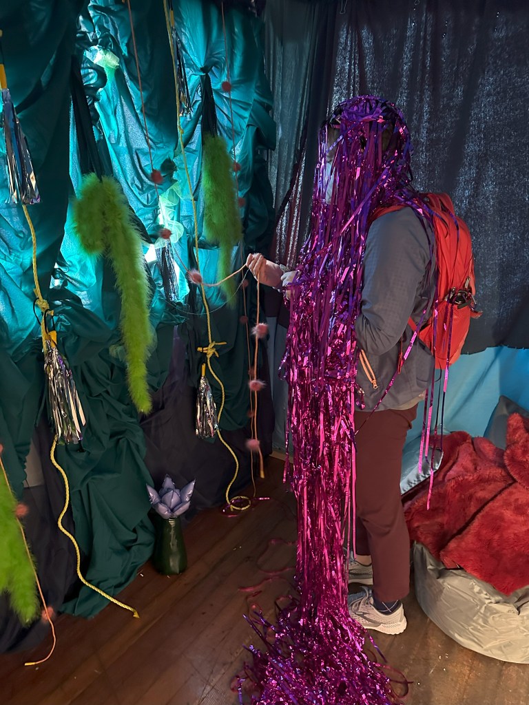 A participant interacts with an artwork by moira williams. The participant wears a gray jacket, reddish-brown pants, and a red backpack. Shimmering purple tinsel almost completely covers their head and torso. They hold a string with small, soft pom-poms in their right hand. The backdrop is a dynamic arrangement of teal and dark-green fabric draped to create a sense of depth and texture. Several green feather boas and shiny silver tassels are suspended from straps cords. There is a green vase holding a purple lotus-shaped object on the wood floor in front of the visitor. To the right of the tinsel-covered person, there's a bit of blue fabric and a reddish-brown pillow or cushion, suggesting seating or further decoration in the space.