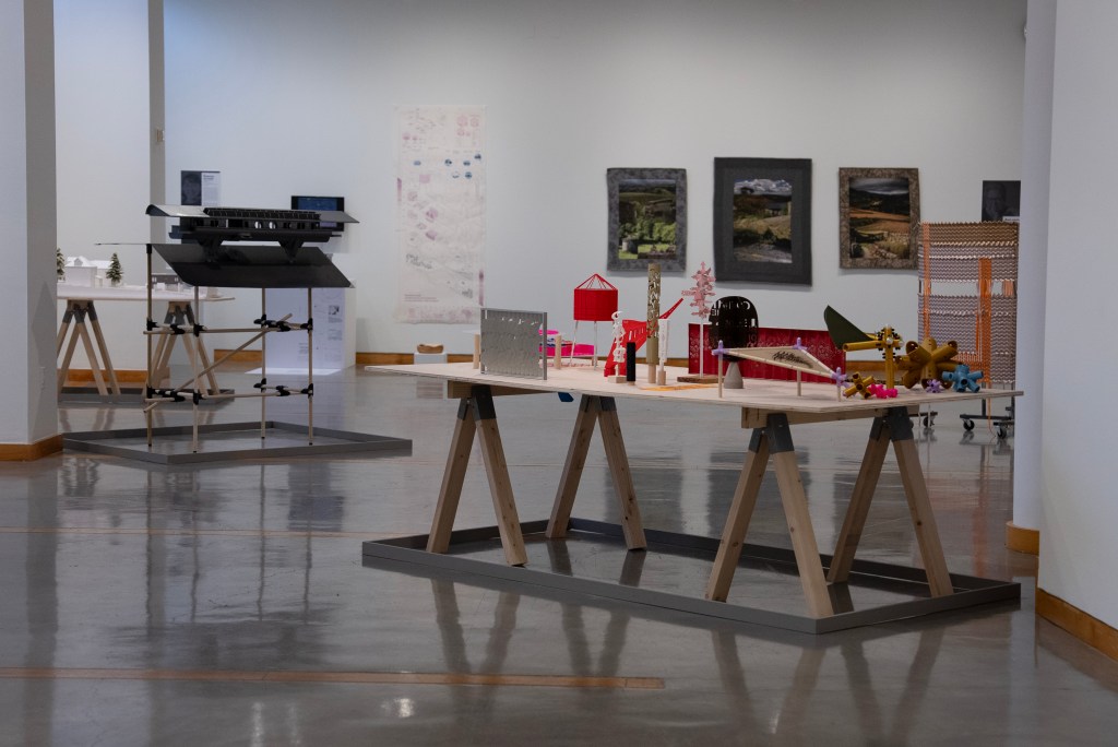 A view into a museum gallery. There are no people in the image. There are many small objects arranged on a plywood table with sawhorse legs in the foreground. On the floor, a can-detectable rail encases the table at the same dimensions as its top. Elsewhere in the gallery, similar detection devices can be seen around the bases of other tables and structures.