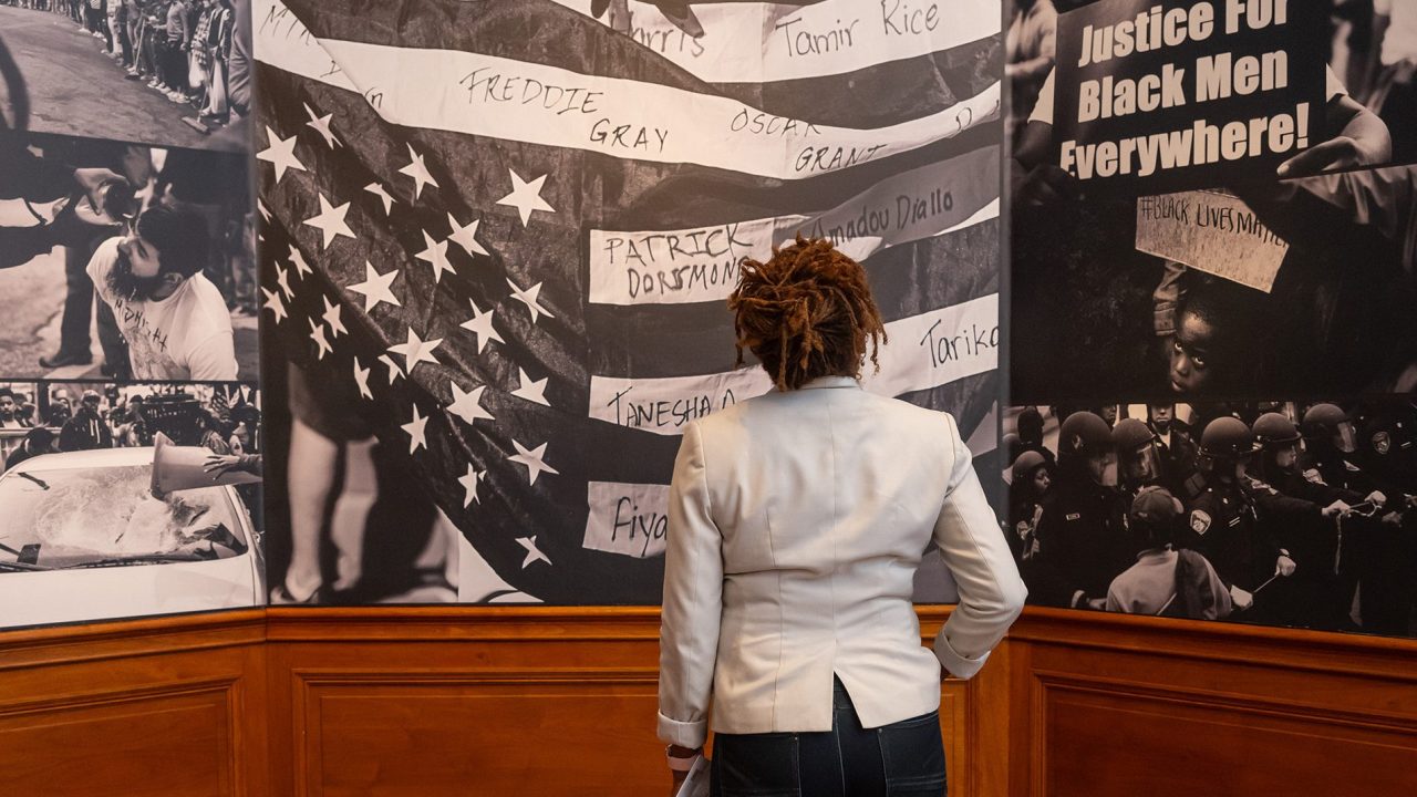 Person standing in a gallery looking at a large mural that includes someone holding up an upside down American flag.