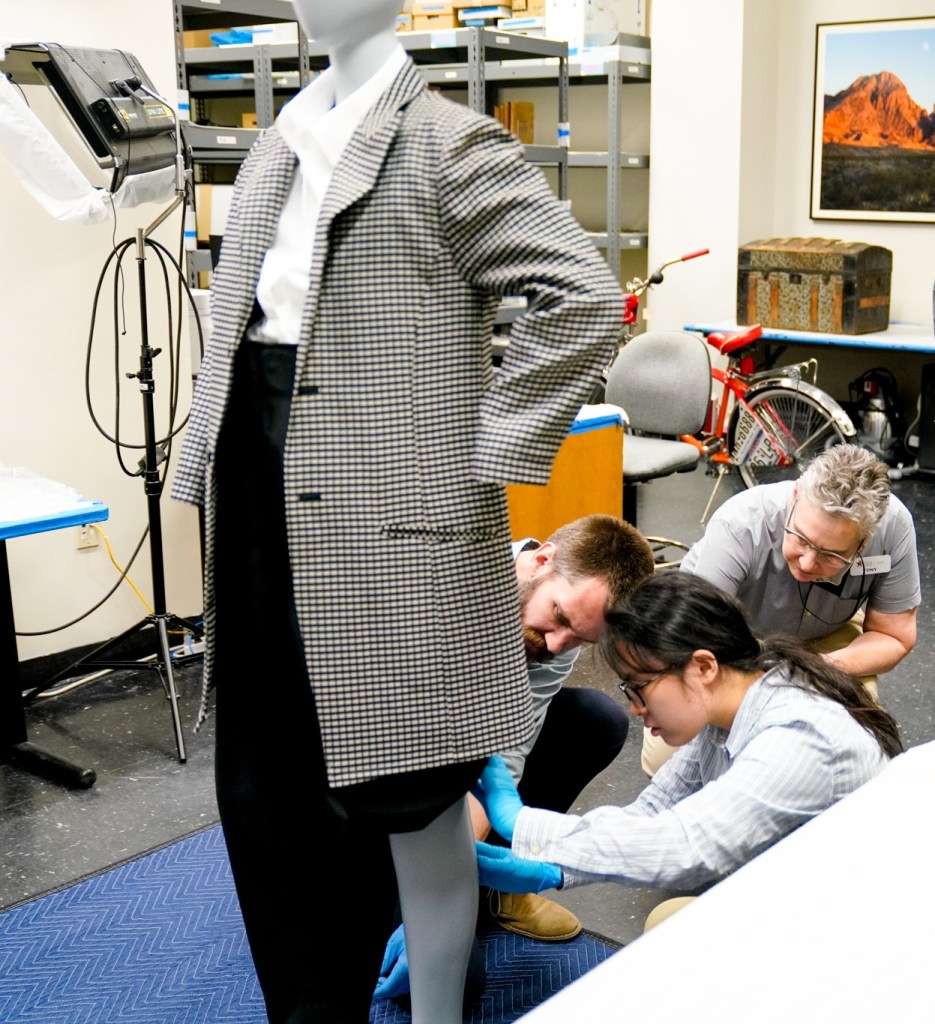 A group of conservators inspecting a mannequin with black pants, a white shirt and long checkered coat