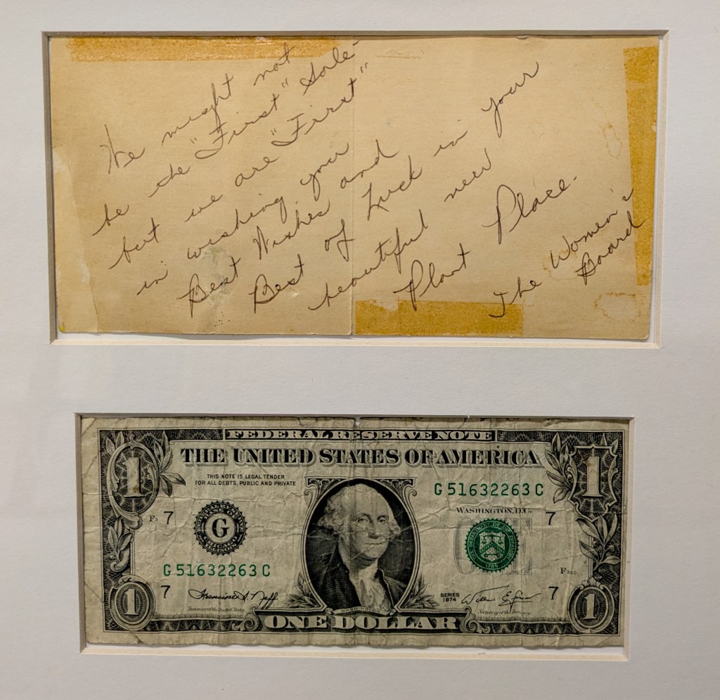 a one dollar bill displayed in a frame with a handwritten note above it.