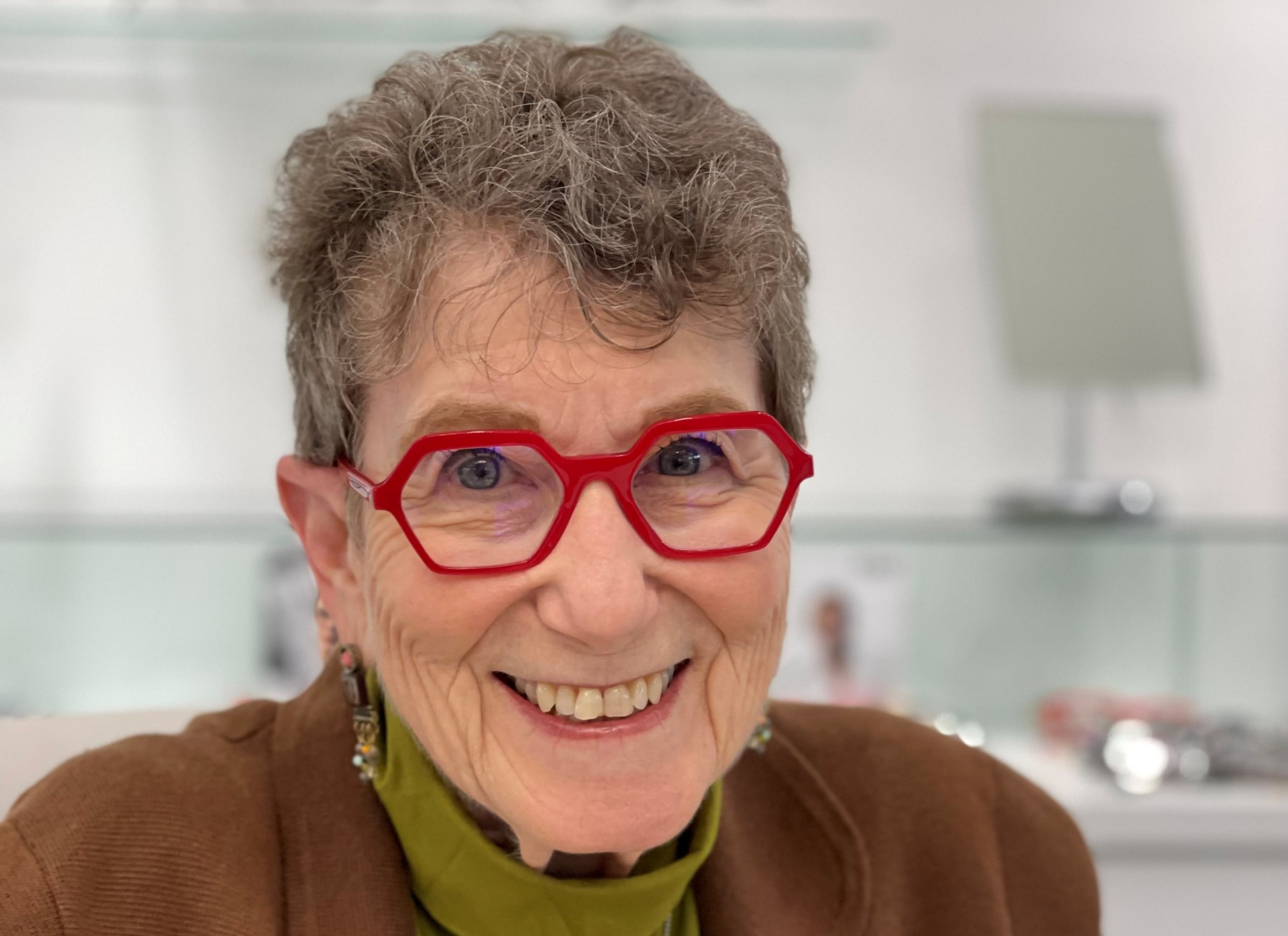 Head shot of a woman with short gray hair wearing red glasses, a green turtleneck shirt and a brown jacket. 