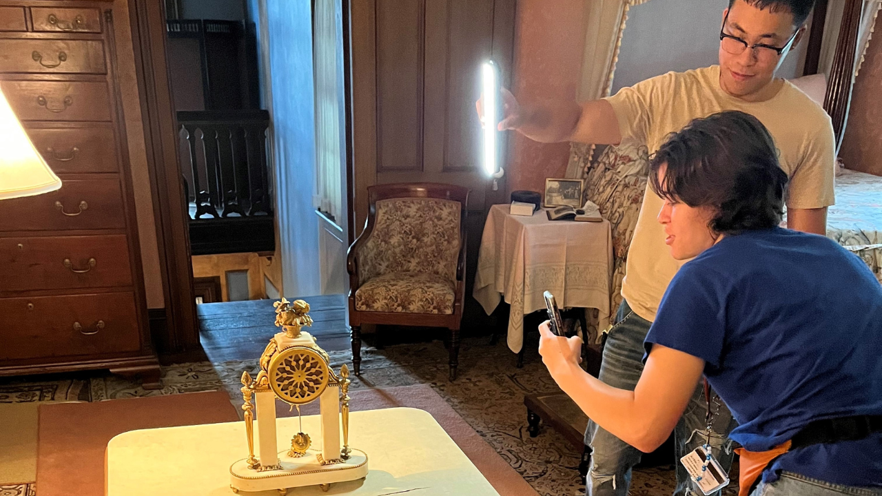 Two people photographing an antique clock on a table, one holding a camera and the other holding a light