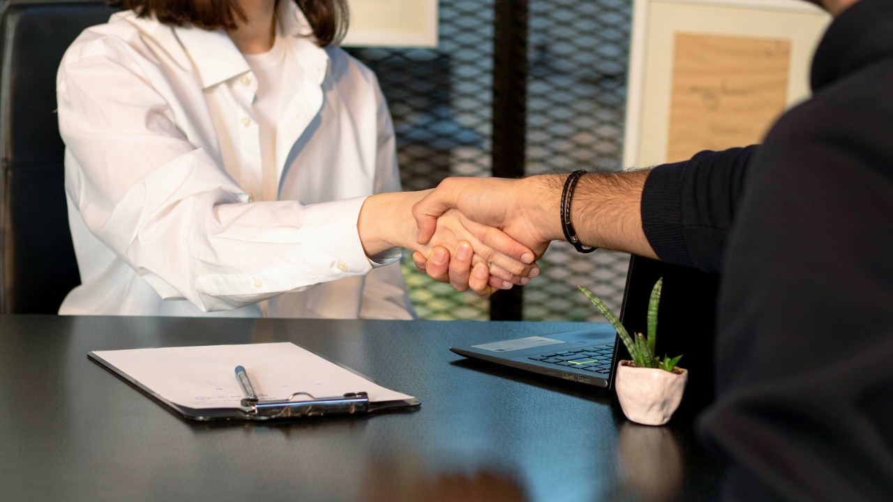 Two people sit across the table from each other with a clipboard and pen in between shaking hands.