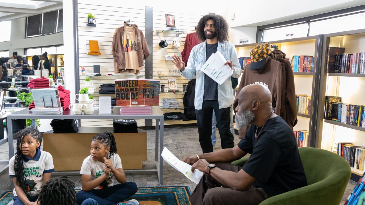 A black male with curly hair enthusiastically speaks to a group seated on a rug in a bookstore. Another person sits nearby, reading a paper. Shelves display books, including Maya Angelou's works. Apparel is visible on the wall.