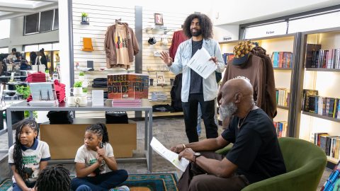 A black male with curly hair enthusiastically speaks to a group seated on a rug in a bookstore. Another person sits nearby, reading a paper. Shelves display books, including Maya Angelou's works. Apparel is visible on the wall.