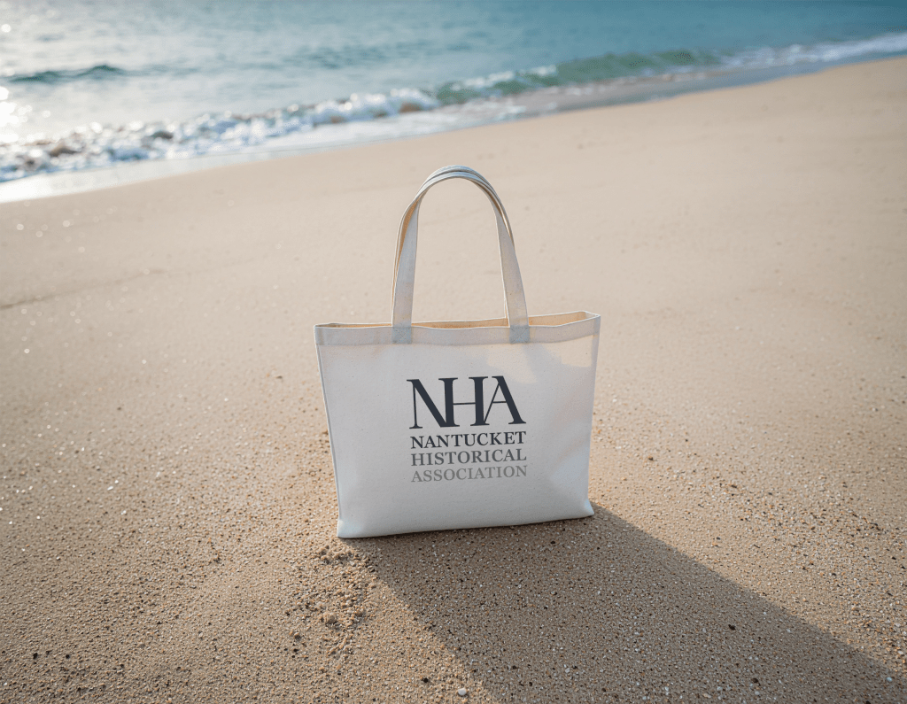 A tote bag on the beach with a logo reading "NHA / Nantucket Historical Association."