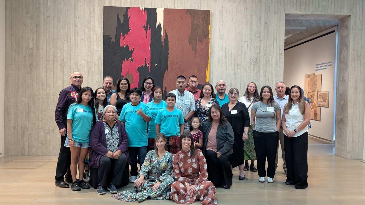 A group of people of all different ages stand and sit in front of a large art piece in a museum gallery.