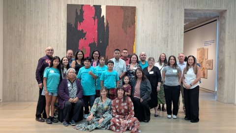 A group of people of all different ages stand and sit in front of a large art piece in a museum gallery.