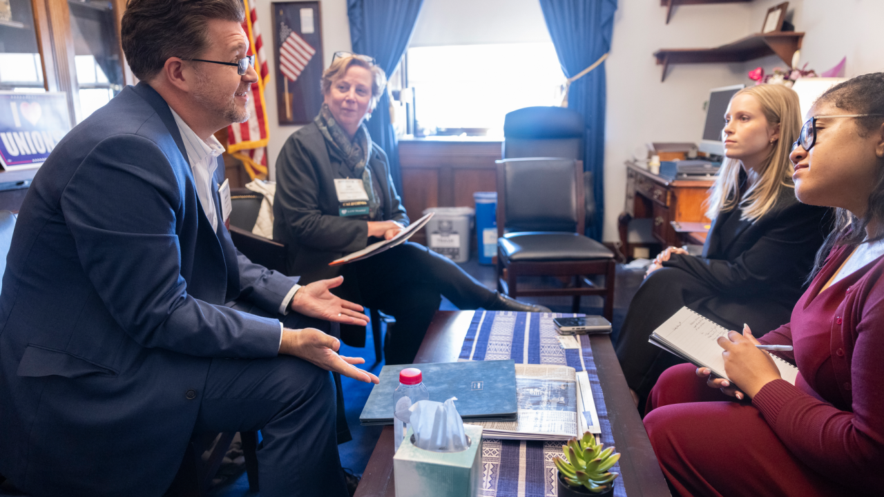 A group of people are sitting in an office setting, engaged in conversation. One person is speaking and gesturing with hands while the others listen attentively and take notes. An American flag is visible in the background, and the room features a desk, bookshelves, and a window with curtains. A small plant, tissues, and other items are on the table between them.