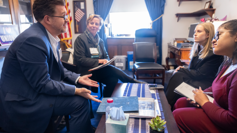 A group of people are sitting in an office setting, engaged in conversation. One person is speaking and gesturing with hands while the others listen attentively and take notes. An American flag is visible in the background, and the room features a desk, bookshelves, and a window with curtains. A small plant, tissues, and other items are on the table between them.