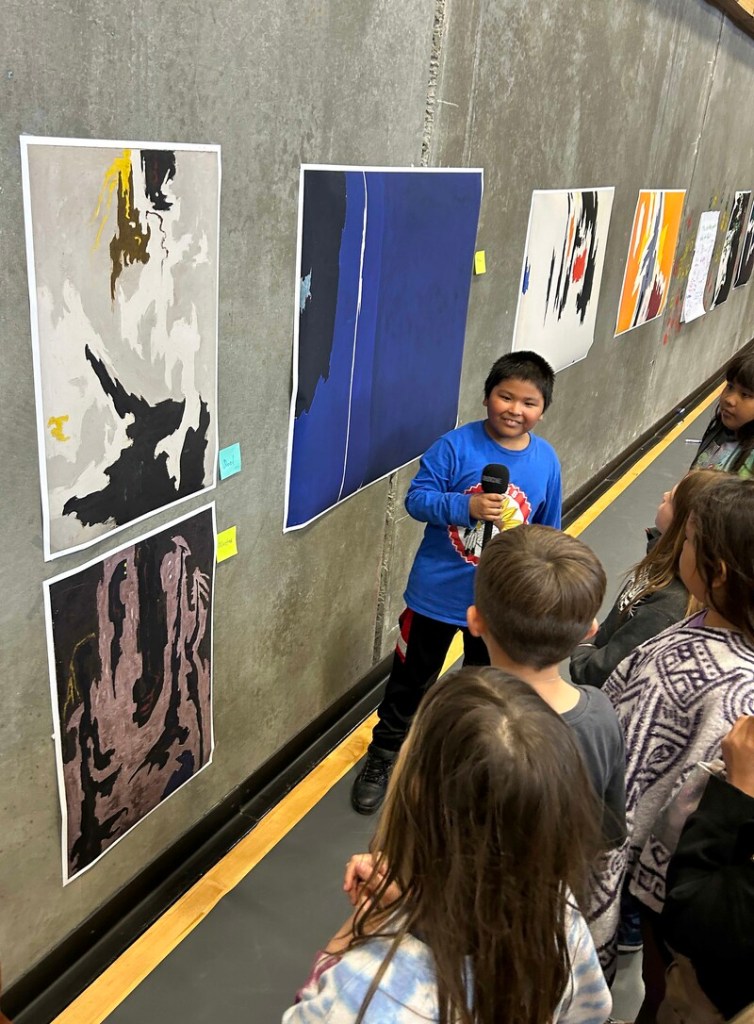 A boy with a microphone stands with his back to printed artwork while speaking to a group of peers.