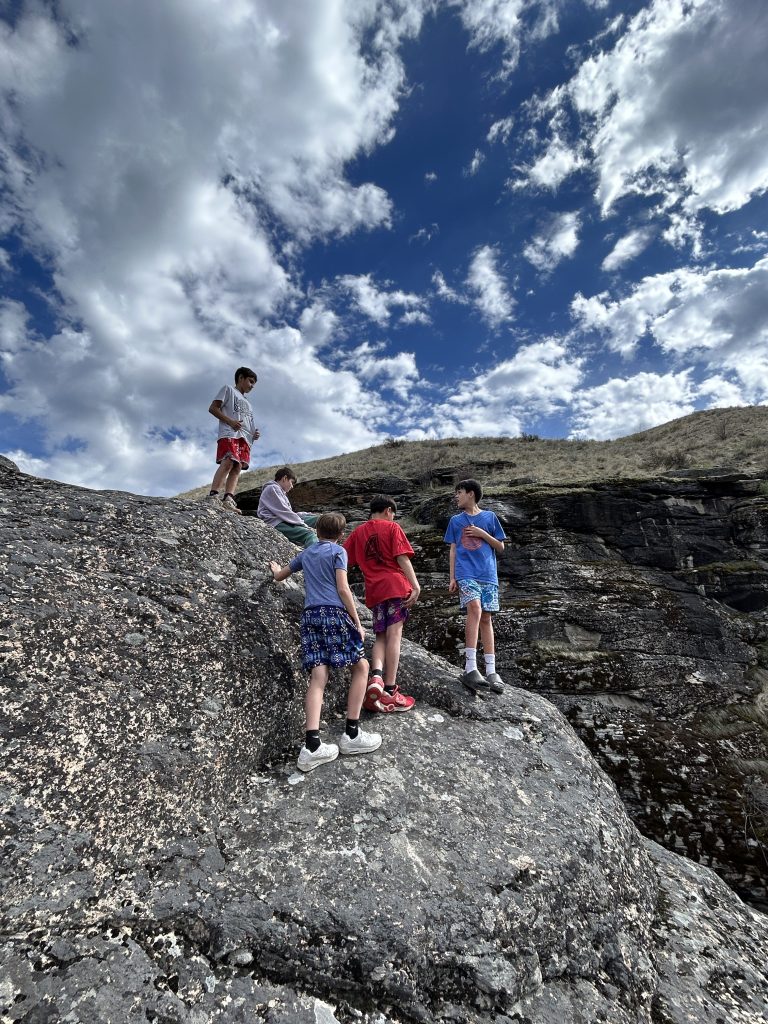Youth standing and sitting on rocky terrain. A blue sky with fluffy clouds is above them.