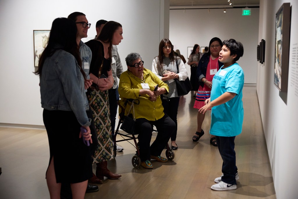 A boy stands in front of an artwork presenting to an intergenerational group of visitors in a gallery.