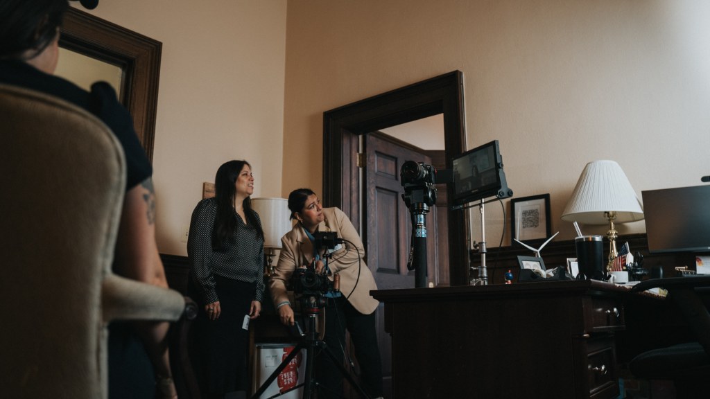 Two women set up a camera and monitor for filming in an office with a desk and computer.