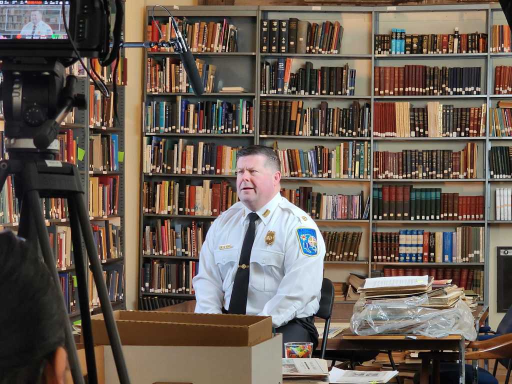 A uniformed fire official sits for an interview in a library surrounded by books and filming equipment.