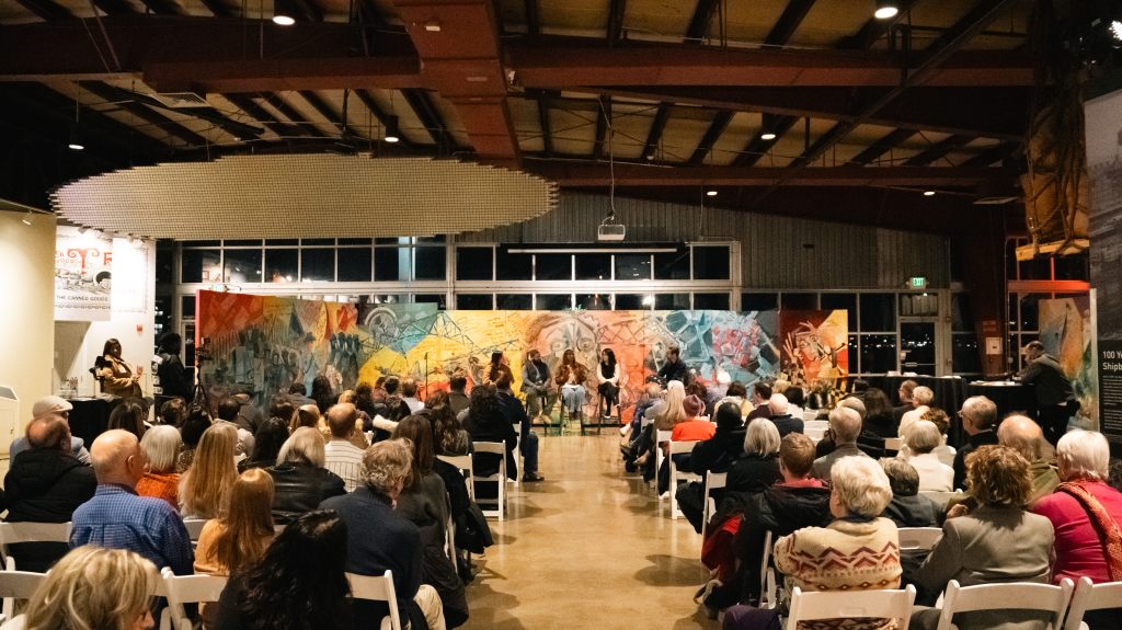 A large audience watches a panel discussion inside a museum, with speakers seated on stools before a vivid, multi-panel mural backdrop.