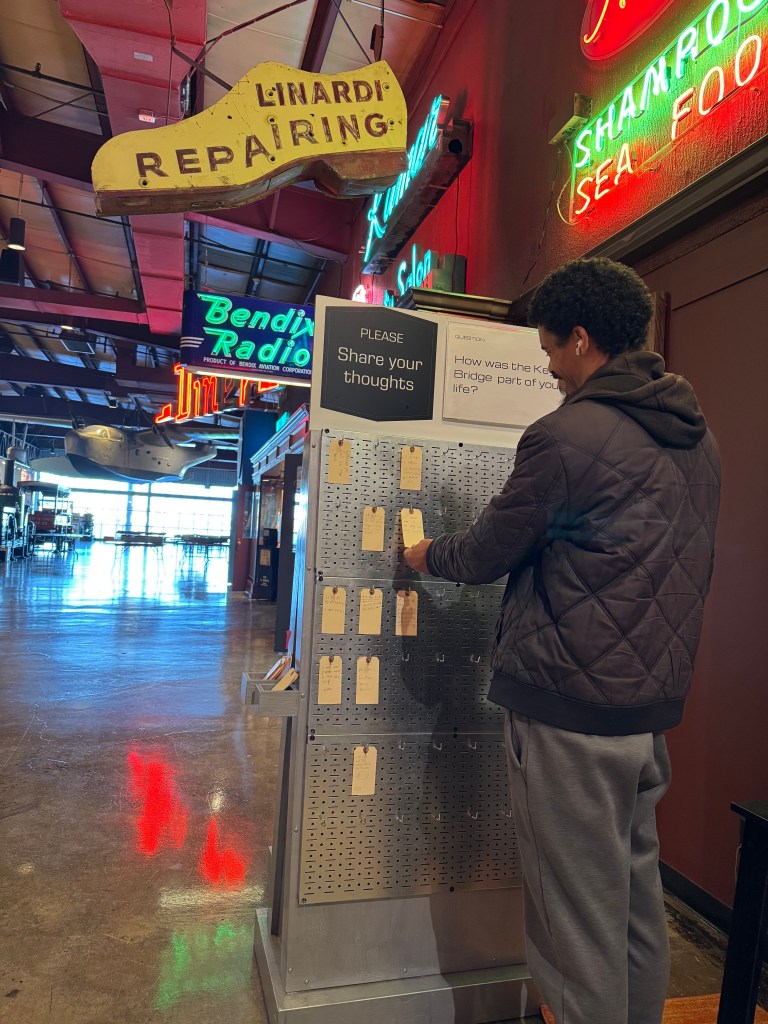 A person stands in front of an interactive display reading a tag written in response to a question asking how the Key Bridge was part of their life.