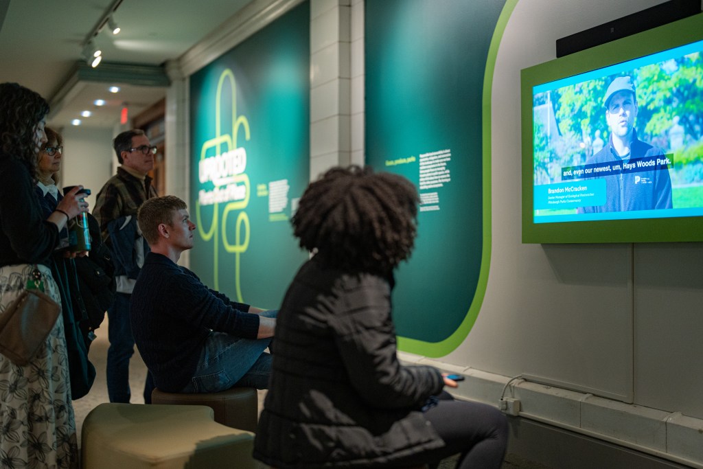 Two seated and three standing visitors watch a video monitor hung on an exhibition wall with the show’s title visible in the background. On the monitor, a man is visible who appears to be speaking to the camera.