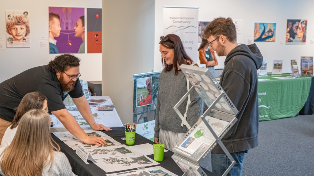 Two people stand in front of a table with pictures of plants and plant specimens on it. Three people are behind the table: two seated women and a standing man, who points at something on the pictures. The room is bright, with other tables and artworks visible along the rear wall.