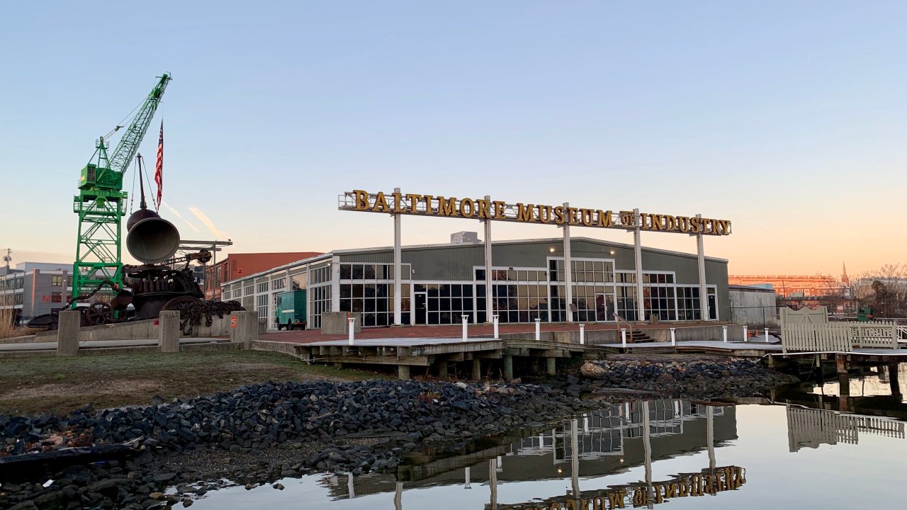 The Baltimore Museum of Industry at sunset, with its sign reflected in calm harbor water and a large green crane nearby.