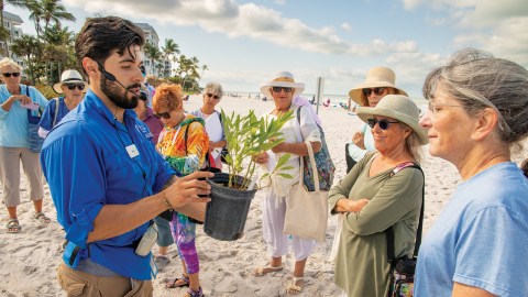 A man holding a potted plant out to a group of people on a beach.