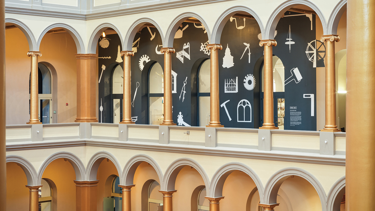 Exterior of a gallery walk with open arches showing black walls behind them with architect's tools painted on them in white.