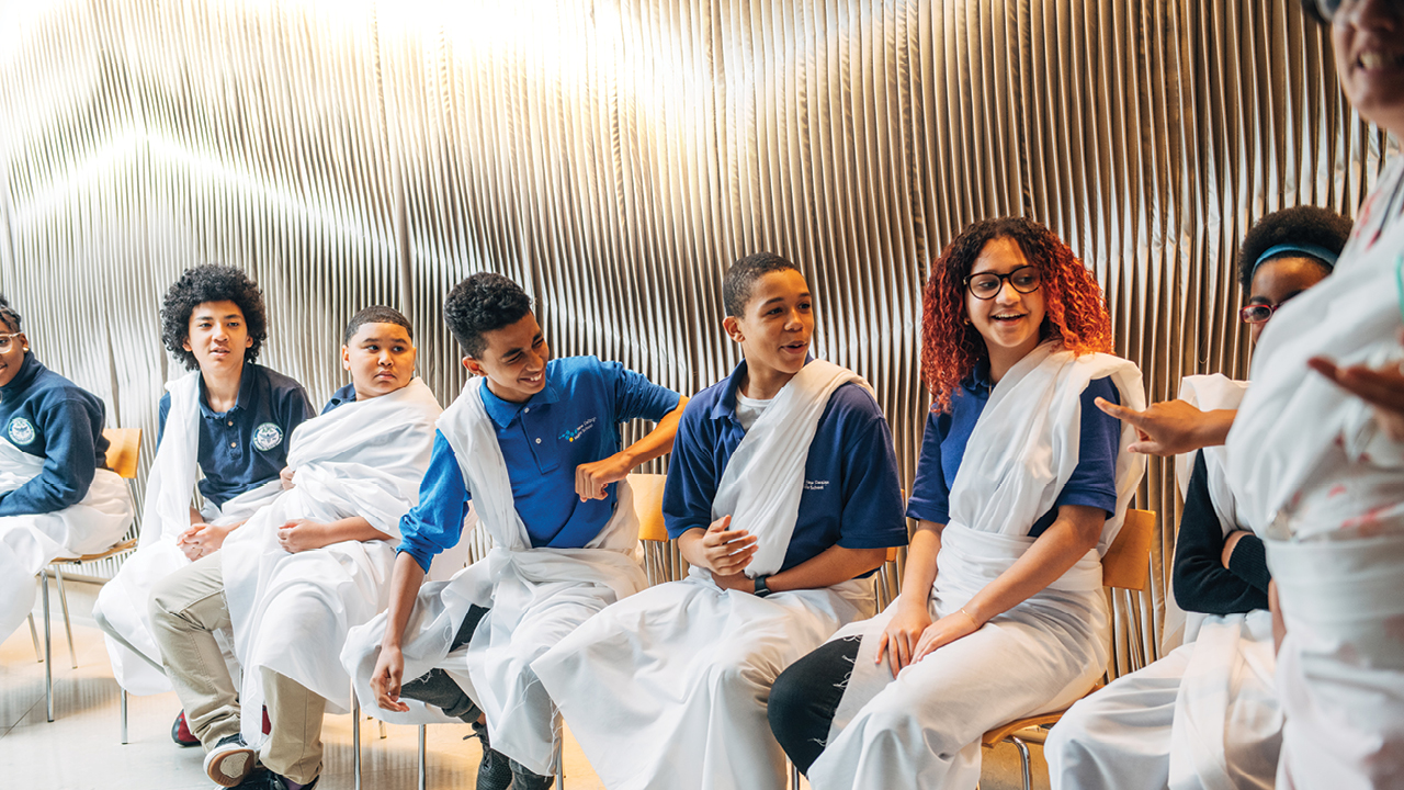 A group of teenagers dressed in white Roman togas and blue t shirts sitting on a bench together