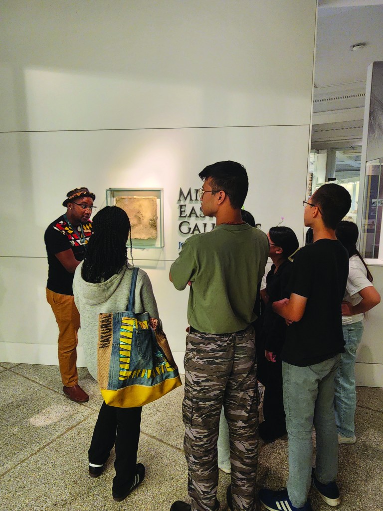 Group of young people standing in a gallery around looking at an object on the wall with a man talking to them.