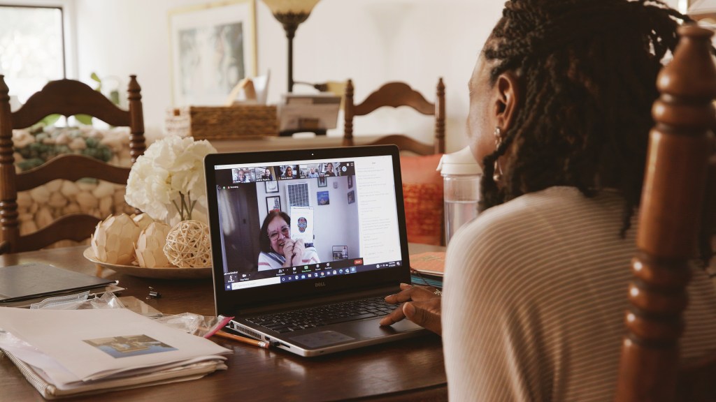 A woman sitting behind a laptop on a table, talking to another woman on her screen.