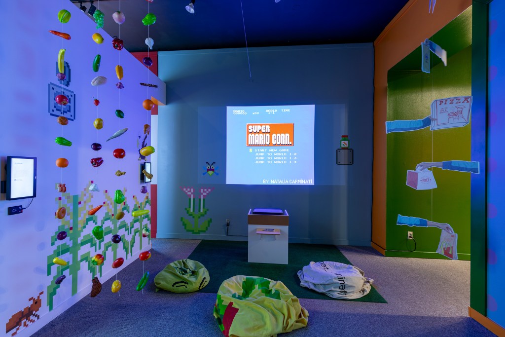 Colorful gallery installation featuring suspended plastic produce, pixelated wall graphics, and a projected title screen for โSuper Mario Corn,โ with beanbag seating and playful food-themed imagery surrounding a central interactive station.