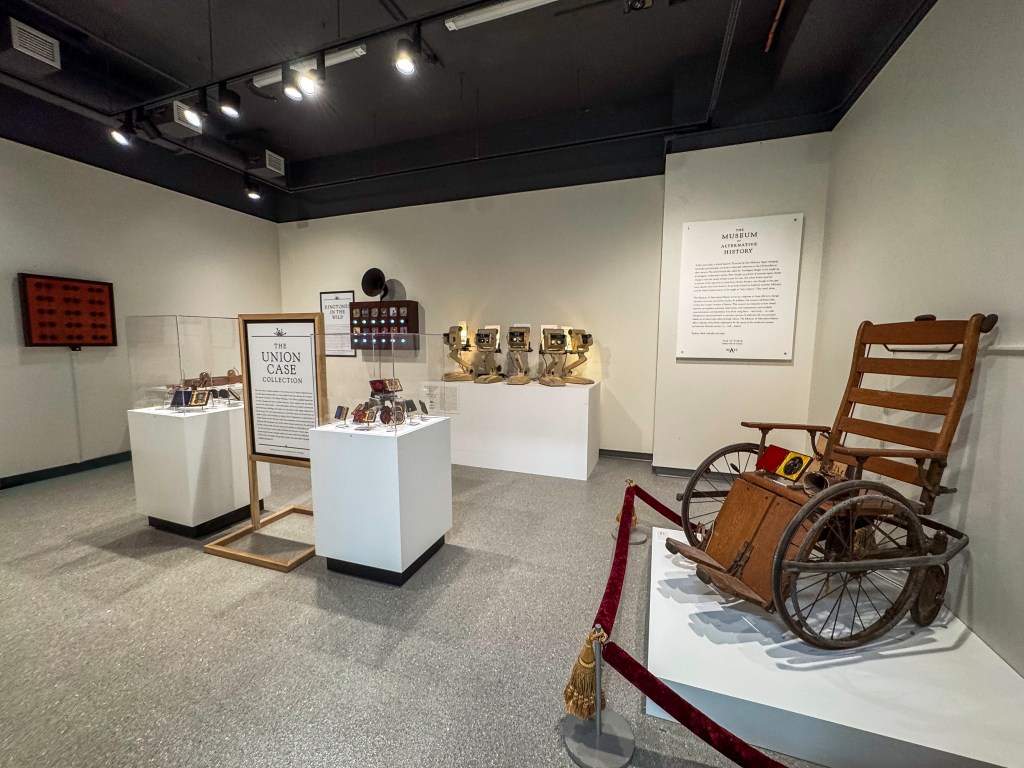 Gallery installation view featuring vintage equipment, framed artifacts in display cases, wall text panels, and an antique wooden wheelchair, representing Tim Guthrieโs Museum of Alternative History during the setup of Blurred Realities.