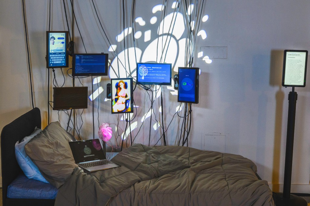 Installation featuring a bed with rumpled blankets, a laptop, and multiple wall-mounted screens displaying chat interfaces, code, and digital figures, with cables cascading downward, representing Chalida Asawakanjanakitโs Romantic Relationships with AI in the Blurred Realities exhibition.