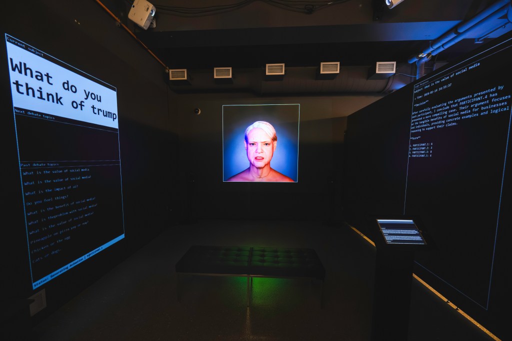 Darkened gallery installation with large wall-mounted text displays showing debate prompts and data, a central illuminated portrait of a human face, and an interactive screen, depicting Calin Segalโs Whispers in the Blurred Realities exhibition.