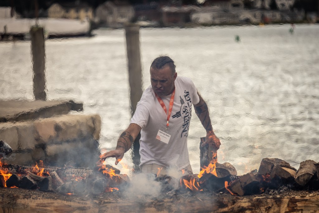 A middle-age Native Mashpee Wampanoag man with long dark-brown hair pulled back with shaved sides, wearing a white t-shirt and a name tag, throws logs onto a dugout canoe on the coast of the Mystic River. The logs on the canoe are on fire, charring the canoe to prepare it to be dug out. The man is wincing at the heat of the flames. 

