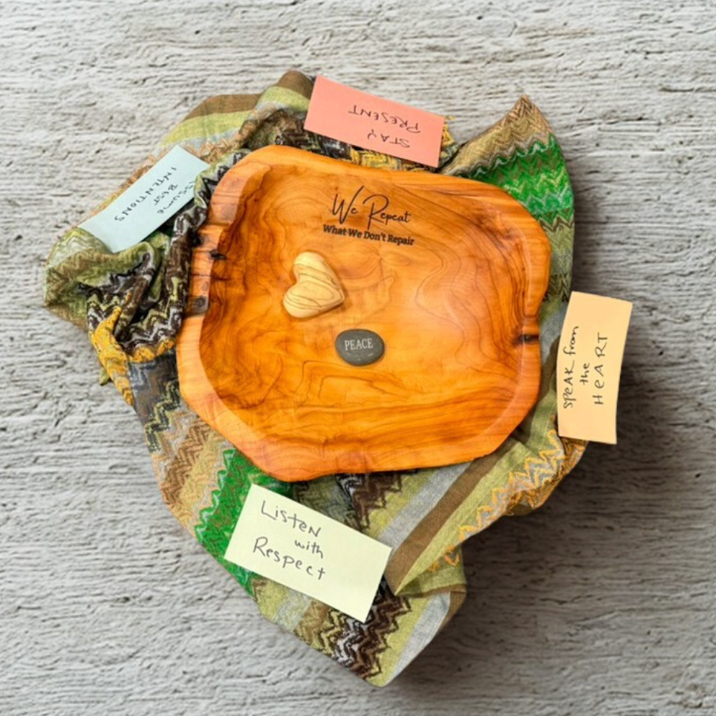 Photograph of a restorative circle centerpiece arranged on patterned fabric, with a wooden bowl engraved with the phrase “We Repeat What We Don’t Repair.” Inside the bowl are small symbolic objects, including a heart-shaped piece and a stone labeled “Peace.” Handwritten cards placed around the fabric read phrases such as “Listen with Respect,” “Speak from the Heart,” “Stay Present,” and “Trust the Process,” representing shared agreements.