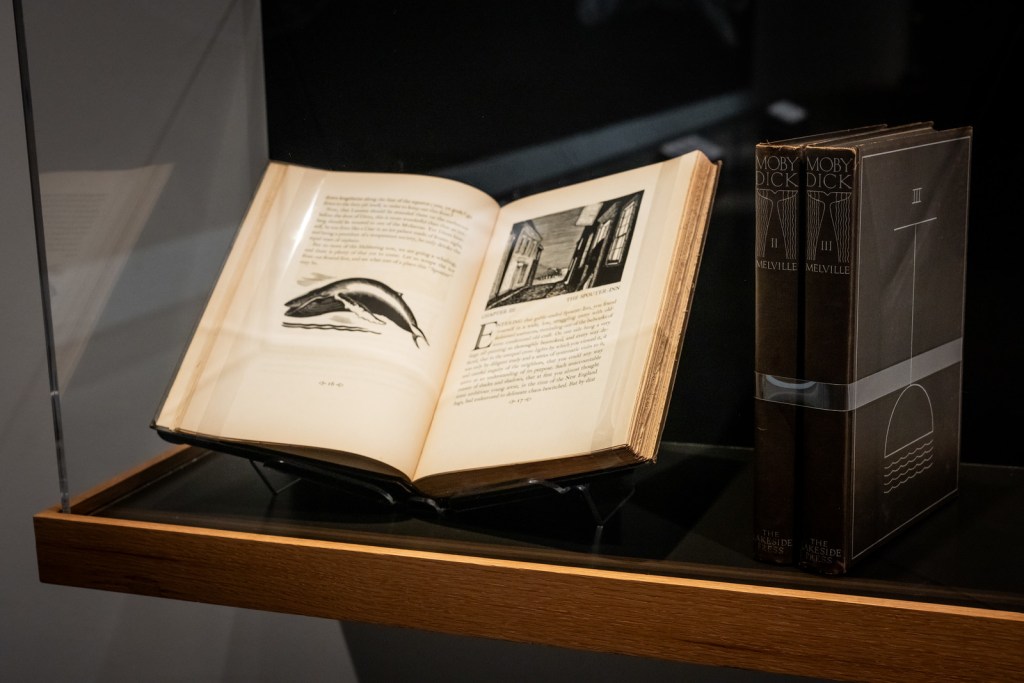 A glass exhibit case shows three books with black covers, which are the three volumes of Rockwell Kent’s illustrated edition of Moby Dick. The first volume is displayed open to the first page of chapter three: The Spouter Inn. The left page shows some text from the end of chapter two with a black-and-white illustration of a humpback whale jumping out of the sea. The right page shows the first page of chapter three with a black and white illustration of an inn by the sea. The other two book volumes are closed and standing upright, as if next to each other on a shelf. 