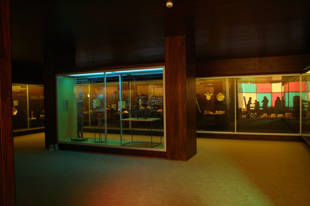 Glass-fronted floor-to-ceiling display cases run along the walls in a square exhibition room with low-lighting. Inside the cases, Timorese objects are mounted on the walls and shelves. In the middle of the room, a floor-to-ceiling glass display case contains three sculptures.